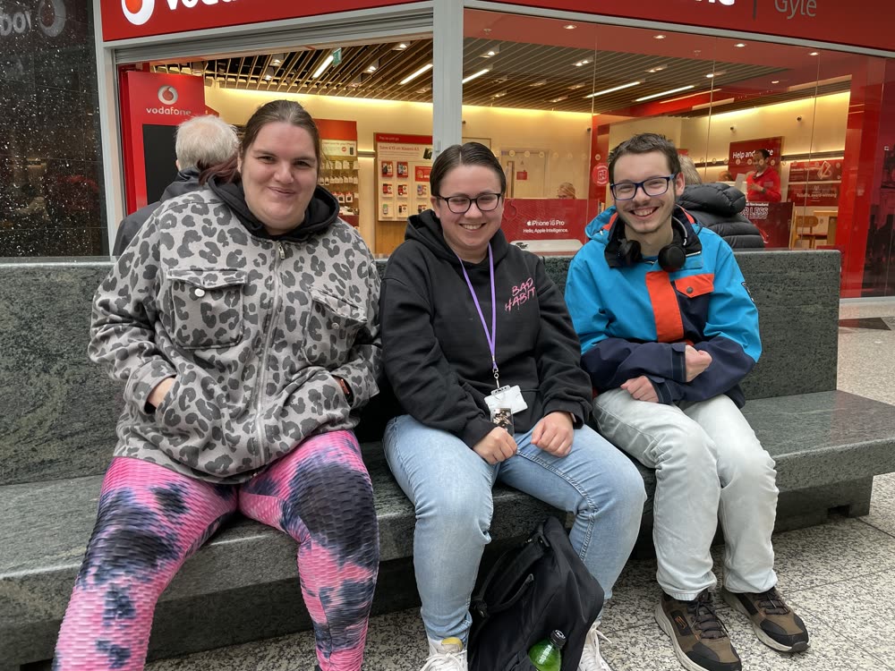 Three friends sitting on a bench at the Gyle centre, smiling at the camera